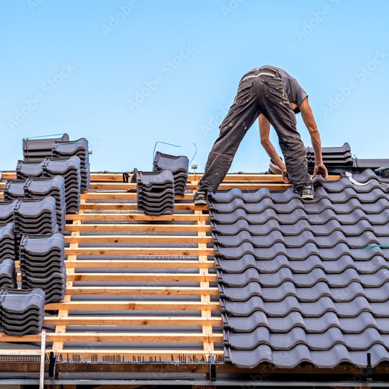 Couvreur installant des tuiles sombres sur un toit en construction, avec des lattes en bois et des piles de tuiles neuves sous un ciel bleu.