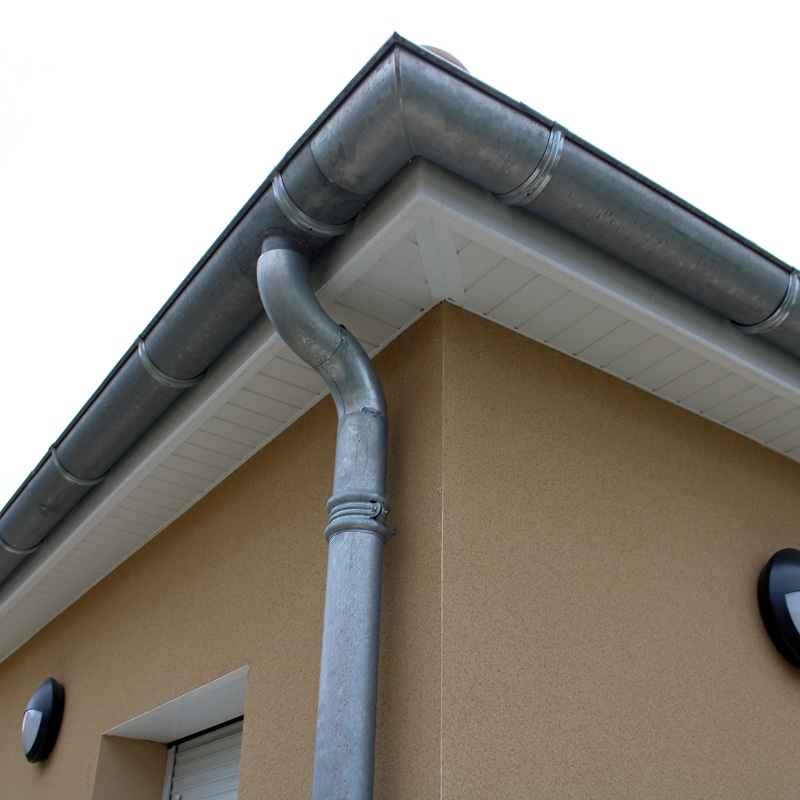 Gouttière et descente d'eau en zinc gris sur le coin d'une maison beige, avec soffite blanc et luminaires extérieurs.