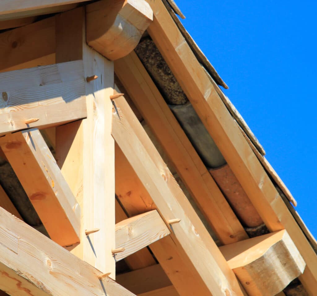 Détail de charpente de toit en bois avec poutres claires et assemblage traditionnel par chevilles apparentes sous ciel bleu.