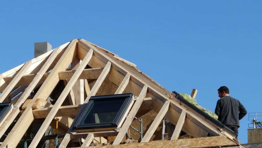 Charpente de toit en bois en construction avec deux fenêtres de toit. Un ouvrier inspecte l'installation sous un ciel bleu.