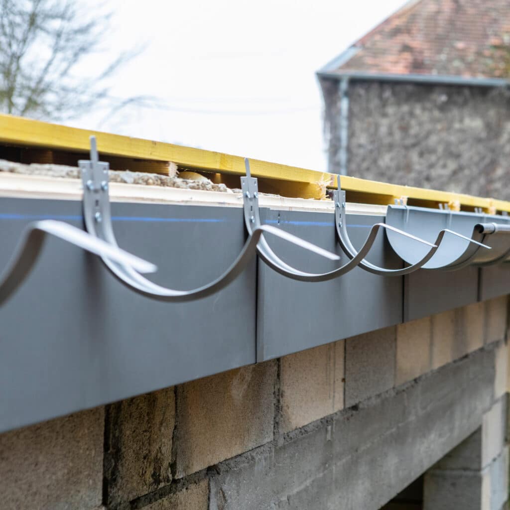 Gouttière grise et ses supports incurvés installés sur une toiture en construction. Charpente en bois jaune au-dessus, mur en blocs de béton.