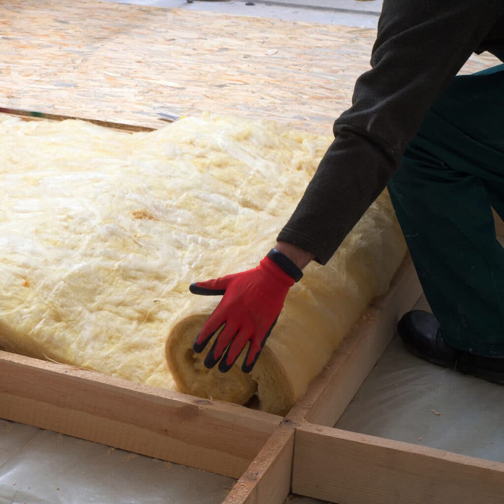 Ouvrier en gants rouges déroule un rouleau de laine minérale entre des poutres en bois, isolant un plancher en construction.