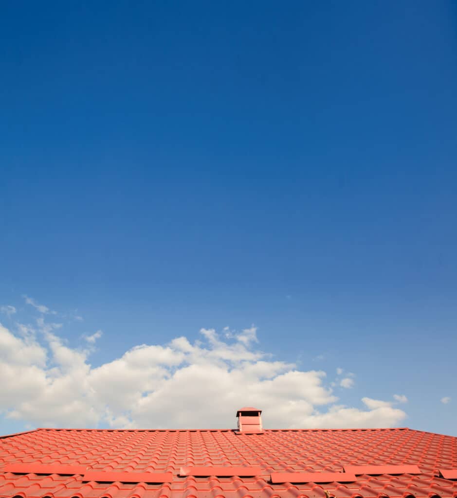 Toit de maison en tuiles rouges brillantes sous un ciel bleu lumineux avec quelques nuages blancs. Une cheminée rouge se dresse sur le toit.