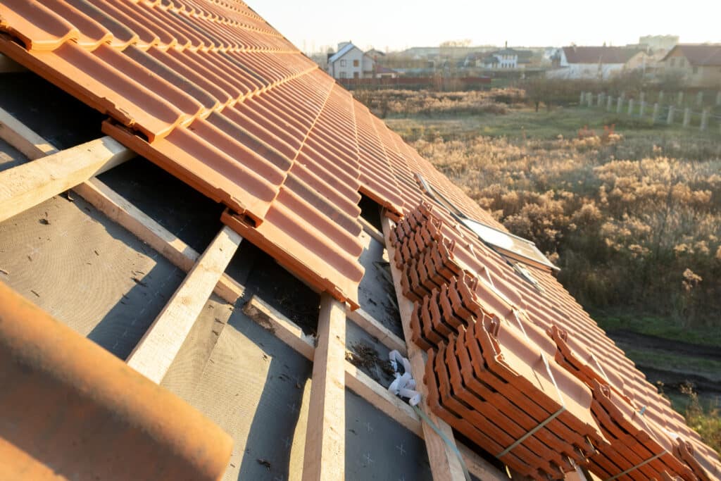 Toiture en construction avec tuiles en terre cuite posées et empilées, charpente bois et membrane d'étanchéité visibles. Arrière-plan de maisons et champs au lever/coucher du soleil.